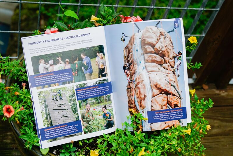 The image shows an open magazine surrounded by greenery and flowers. The magazine features a collage of photos related to community engagement, including people gathered together and a climbing wall. The right page displays a close-up of the climbing wall, emphasizing its texture and design.